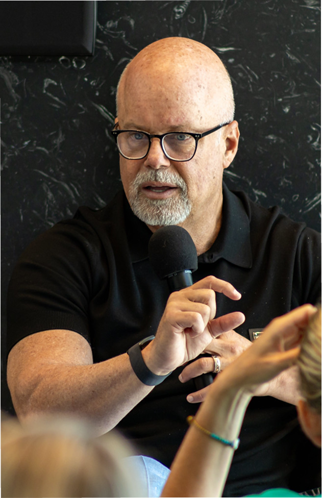 Eric Worre holding a microphone and smiling while standing in front of a dark, textured wall with his name displayed in the corner.