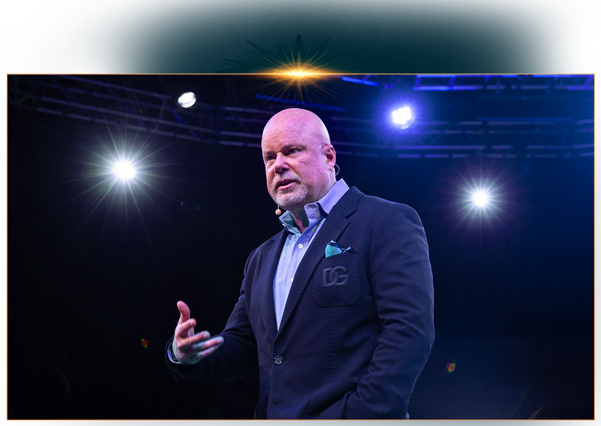 Eric Worre gesturing while speaking on a dark stage illuminated by bright blue and white spotlights.