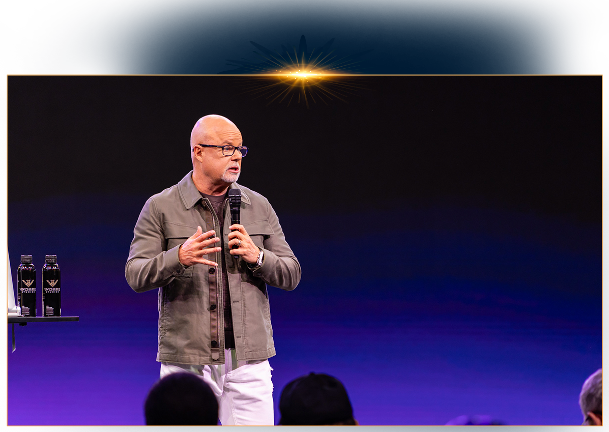 Eric Worre, wearing a green jacket and glasses, speaking into a microphone on a stage with a purple and blue lit background.