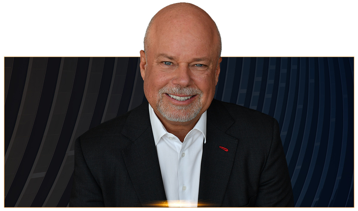 Bald man with a trimmed beard wearing a dark suit and white shirt smiling at the camera against a dark patterned background.
