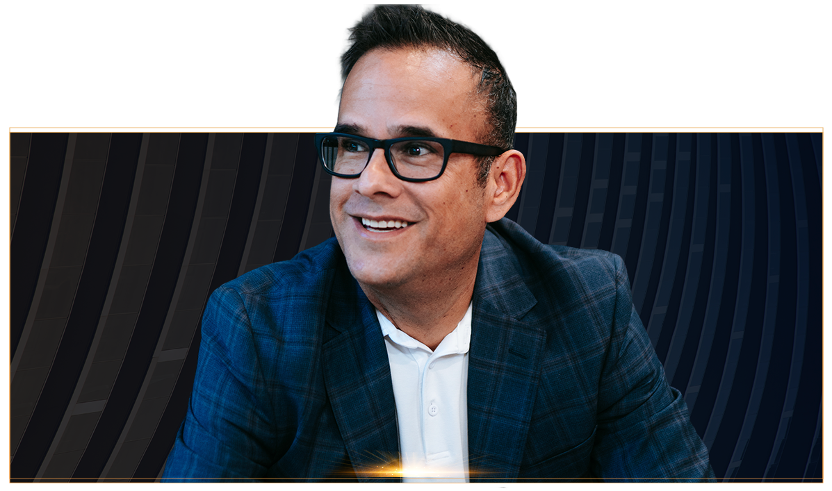 Man wearing glasses and a blue blazer smiling and looking to the side against a dark patterned background.