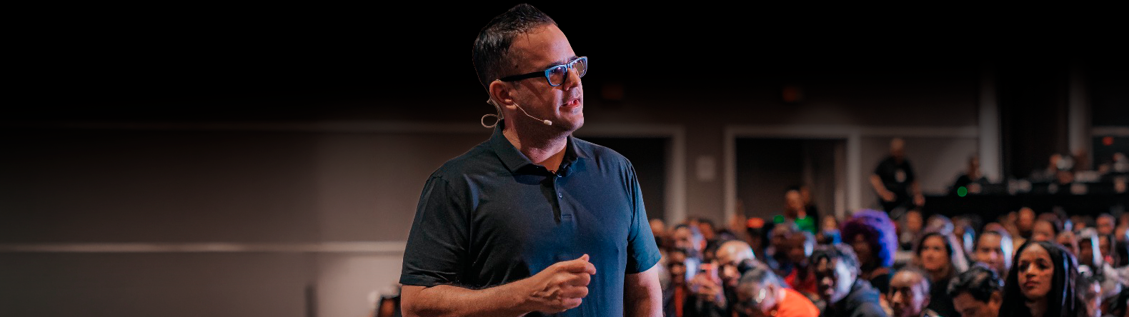 A man with glasses and a headset microphone speaking on stage, with a blurred audience in the background.