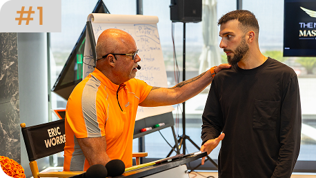 Eric Worre, wearing a bright orange shirt, places a hand on a younger man's shoulder while speaking to him in a studio setting.