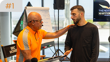Eric Worre, wearing a bright orange shirt, places a hand on a younger man's shoulder while speaking to him in a studio setting.